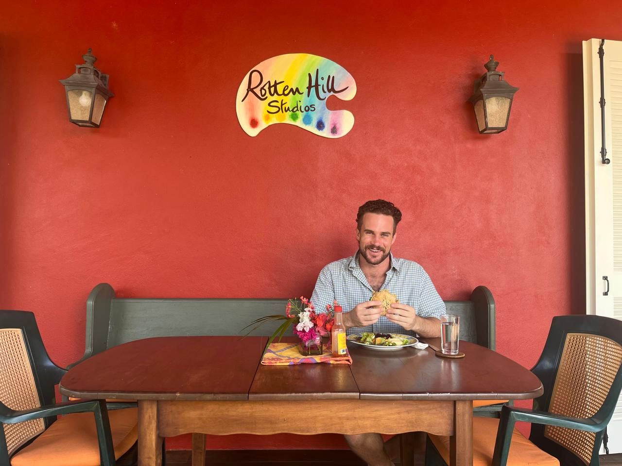 Man dining at Raton Hill Shop restaurant, sitting at wooden table with food and drinks against red wall.