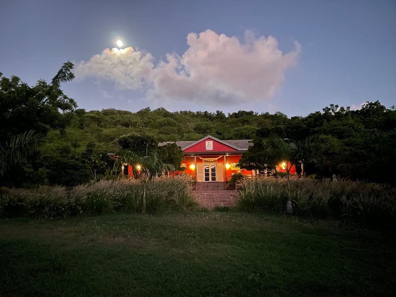 A glowing red farmhouse illuminated at dusk with a full moon rising above lush green trees in a tropical setting.
