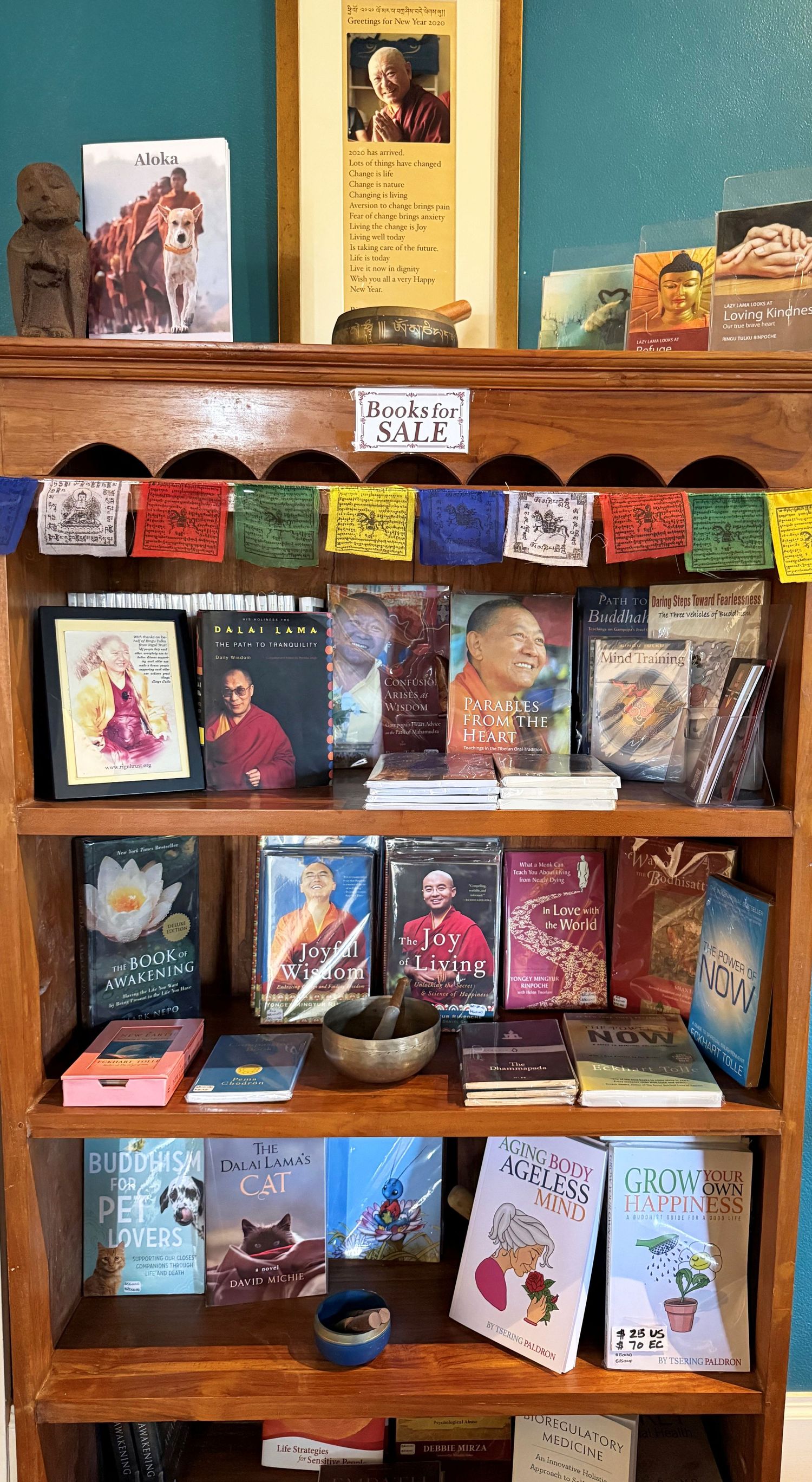 Wooden bookshelf displaying spiritual and Buddhist books for sale, including Dalai Lama titles and Joy of Living.