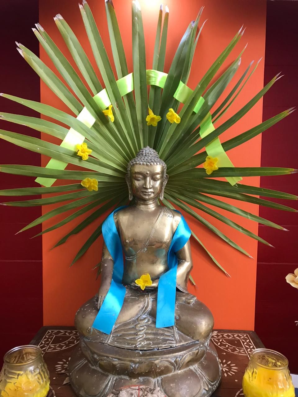 Golden Buddha statue adorned with blue sash, palm leaves, and yellow flowers against a vibrant orange wall.