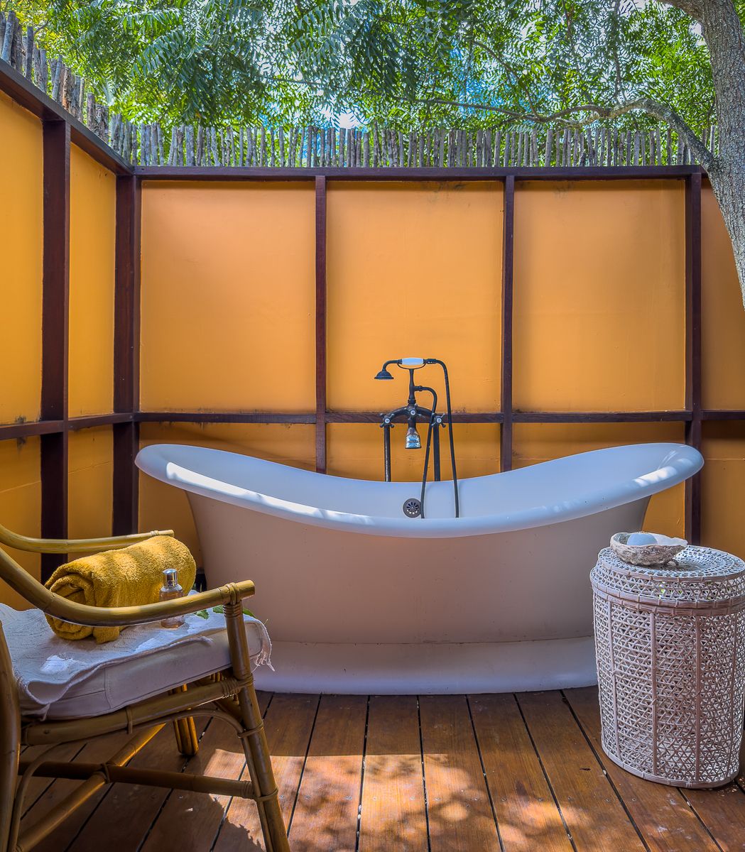 Luxurious freestanding soaking tub on a wooden deck with golden yellow walls and lush greenery in an outdoor bathroom.
