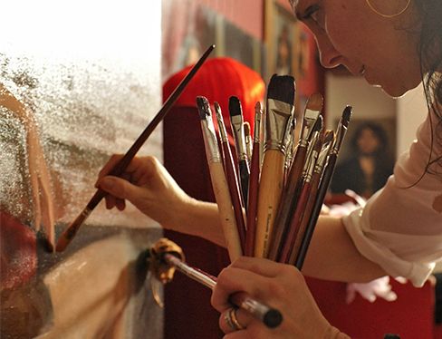 Artist holding multiple paintbrushes while working on a detailed canvas painting in studio.
