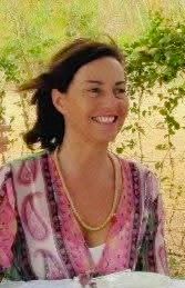 Smiling woman with dark hair wearing a pink patterned top and gold necklace outdoors in natural setting.