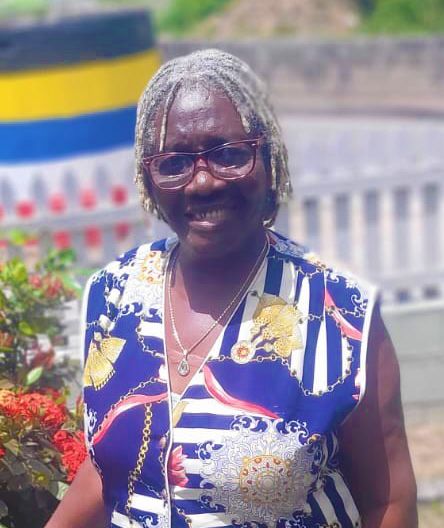 Smiling older woman with gray locs wearing blue floral dress and glasses outdoors near colorful flag.