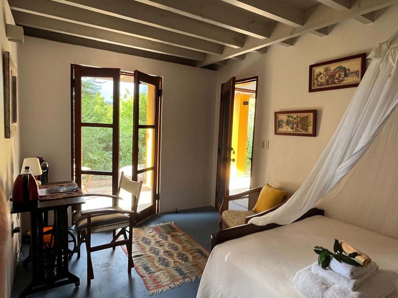 Charming rustic bedroom with canopy bed, wooden beamed ceiling, vintage sewing desk, and French doors opening to a lush garden.