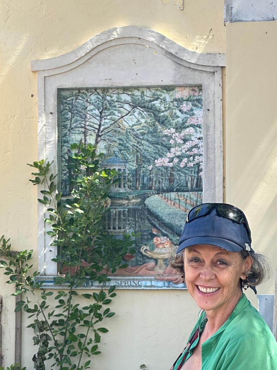 Smiling woman in green shirt and blue cap poses beside an ornate framed garden mural on a yellow wall.