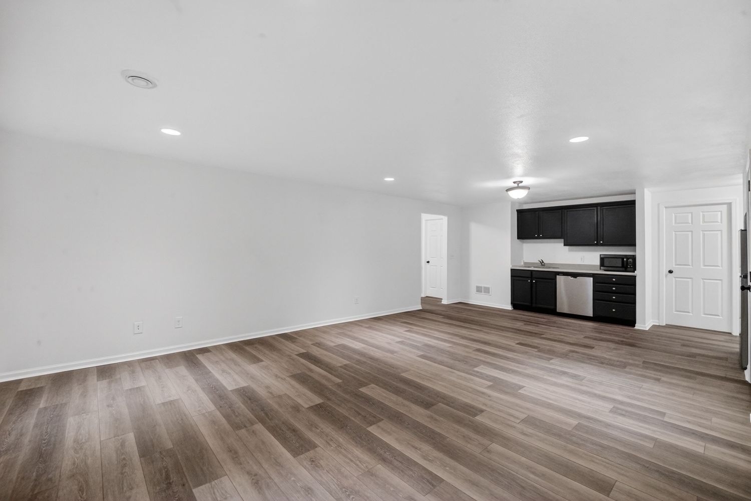 Modern open-concept room with wood-look flooring, white walls, recessed lighting, and black kitchen cabinets.