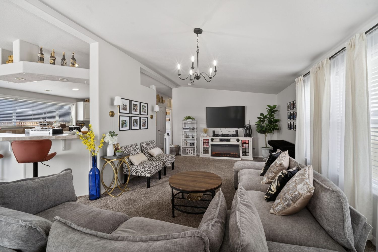 Cozy living room with gray sectional sofa, round wood coffee table, chandelier, TV console, and bright windows with white curtains.
