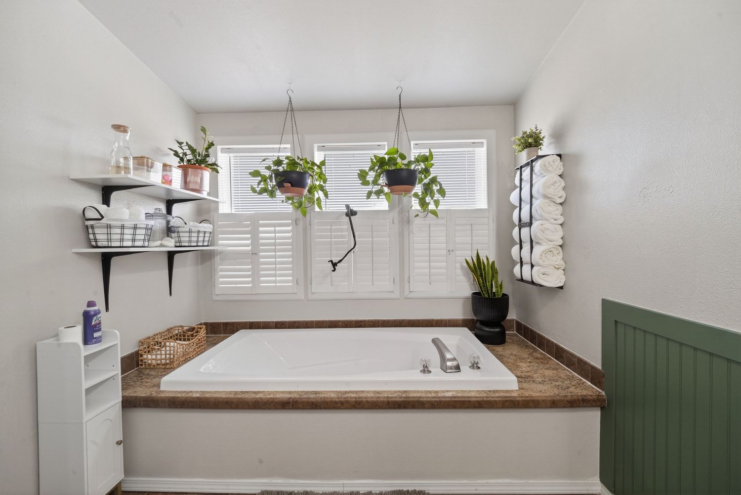 Cozy bathroom with a soaking tub, hanging plants, plantation shutters, open shelving, and green wainscoting accent wall.