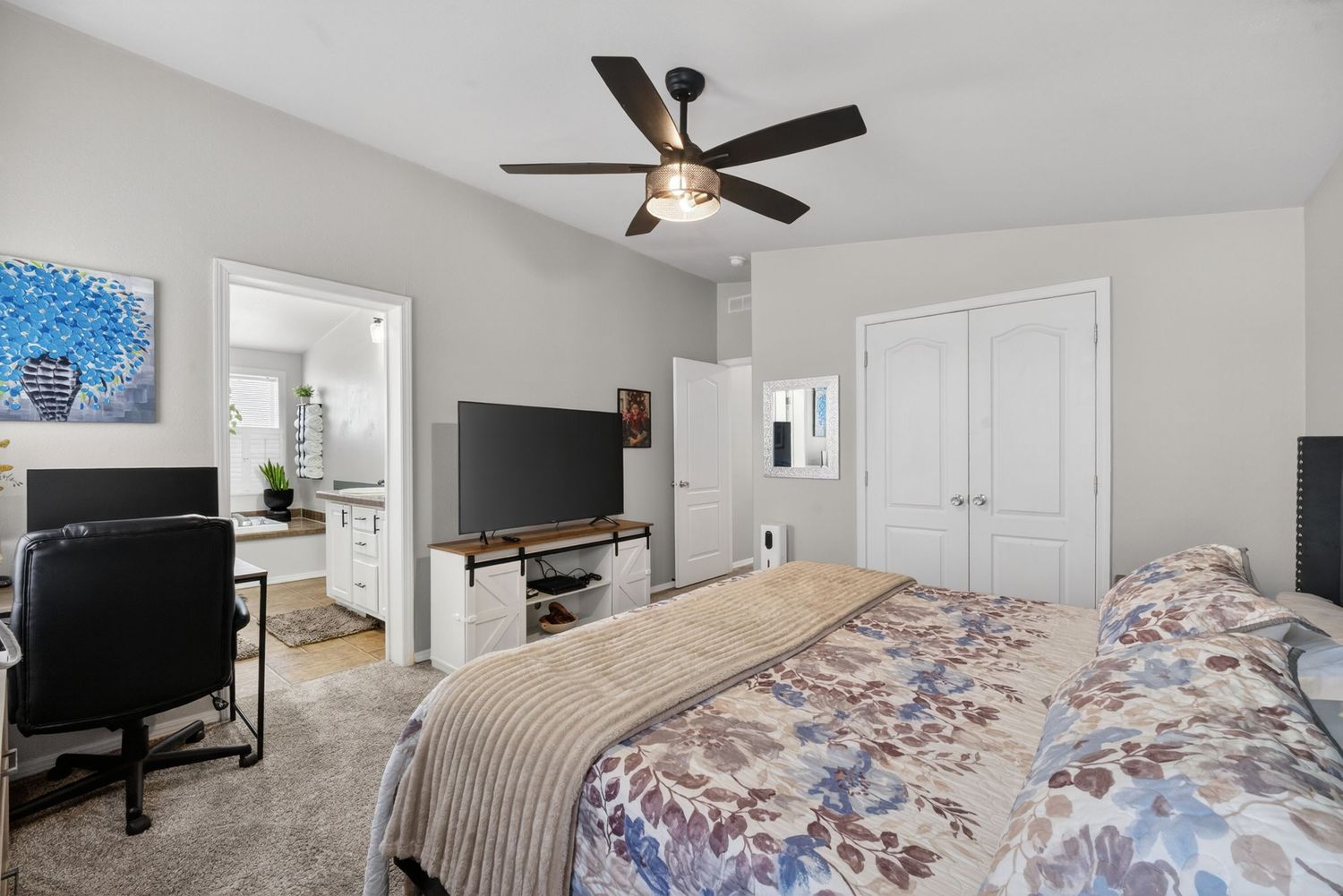Spacious bedroom with ceiling fan, floral bedding, TV, desk area, and double closet doors with neutral carpet flooring.