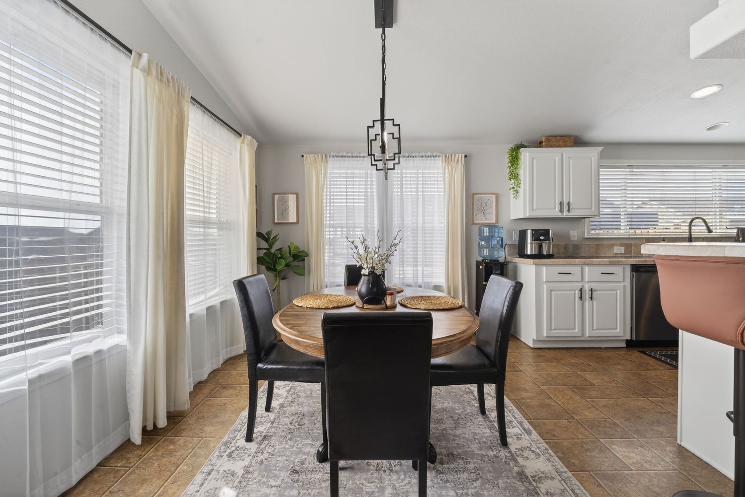 Bright dining room with round wood table, black chairs, chandelier, white kitchen cabinets, and large windows with curtains.