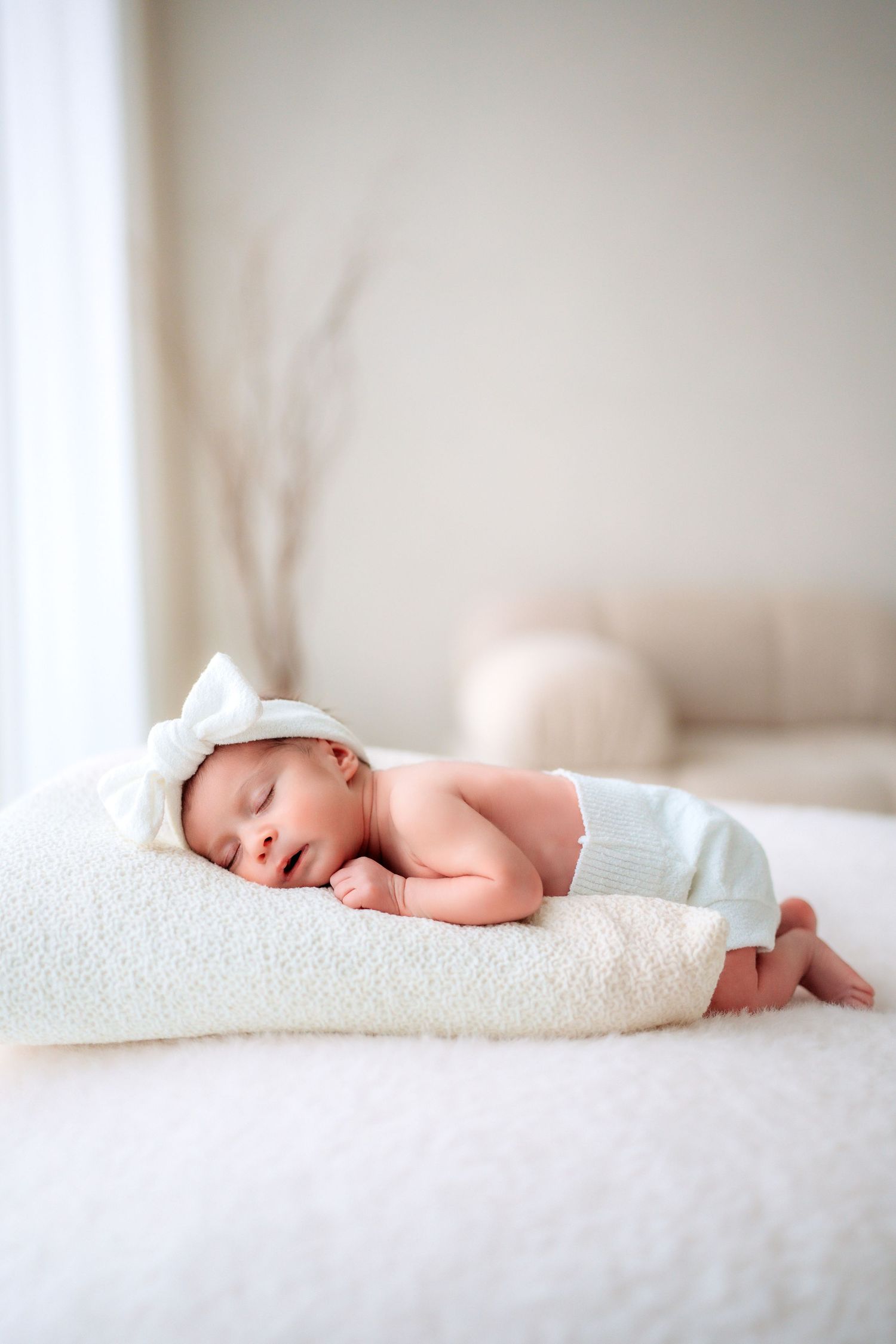 Peaceful newborn baby sleeping on a white pillow wearing a cute knit hat and light blue pants in a bright nursery.