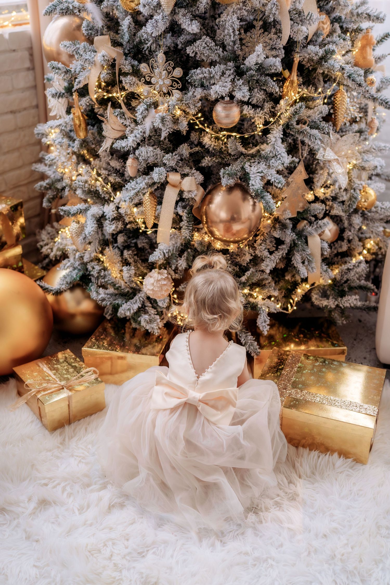 Young girl in elegant white dress with bow sits before decorated Christmas tree with golden ornaments and gift boxes.