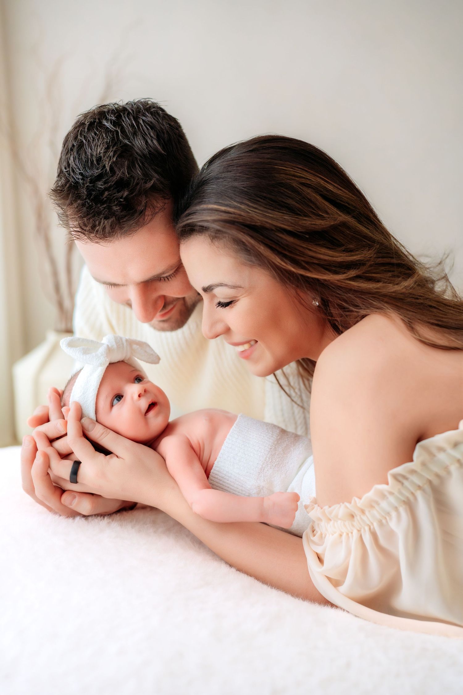 Smiling parents in neutral tones lean over their newborn baby girl wearing a white bow headband.