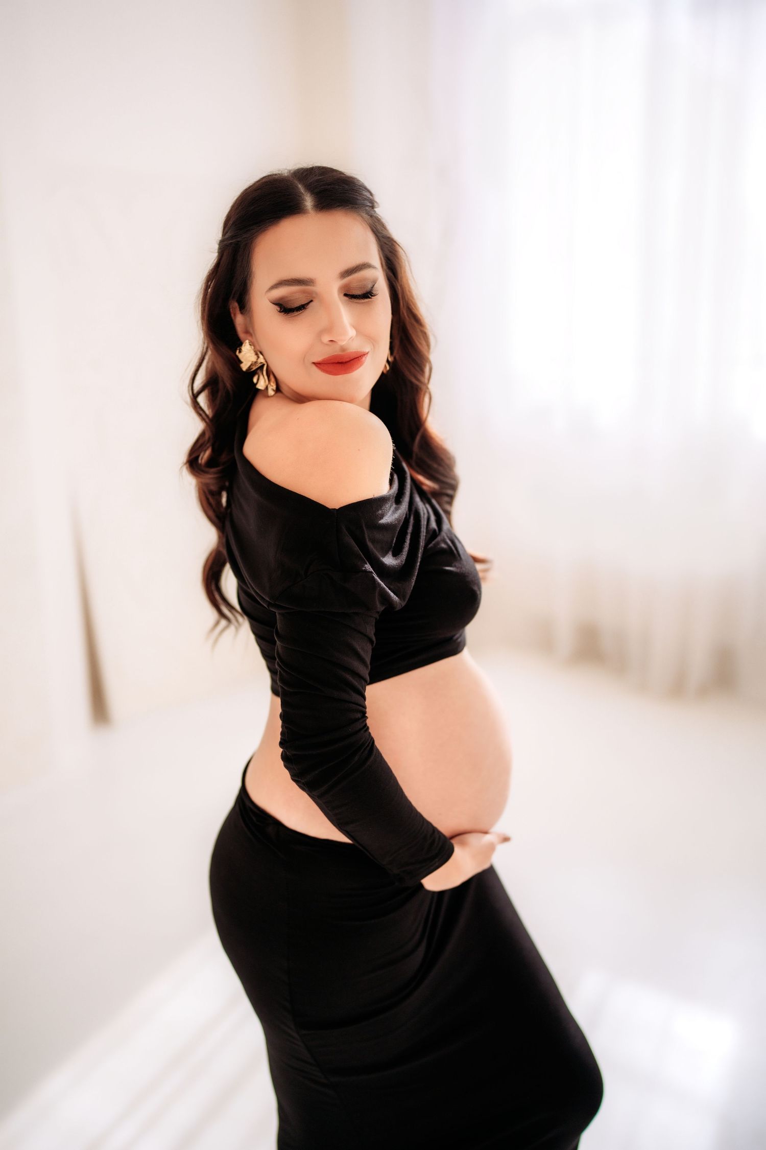 Pregnant woman in black off-shoulder dress cradles baby bump in a bright white room.