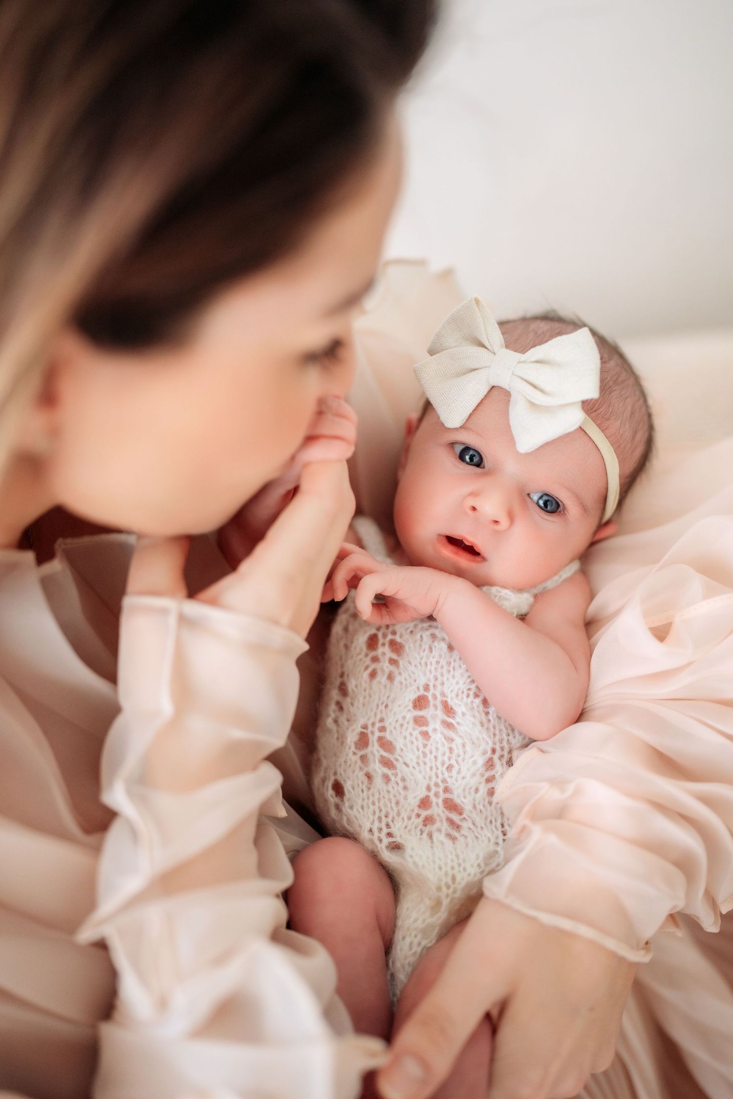 Mother in pink dress kissing newborn baby girls tiny hand while baby looks up with blue eyes.