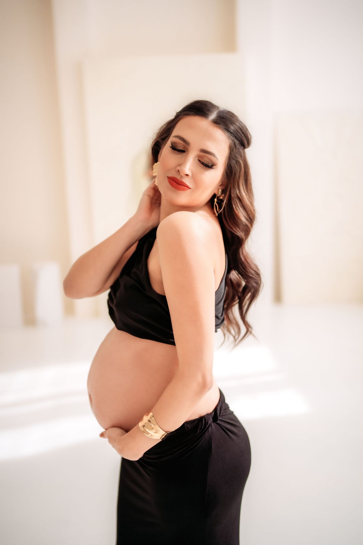 Beautiful pregnant brunette in black outfit with gold bracelet cradles bare baby bump.