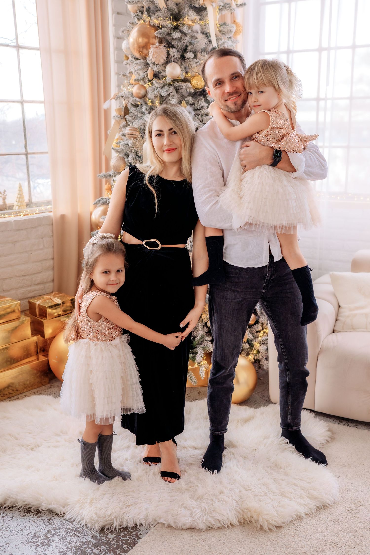Family of four posing by decorated Christmas tree, parents with two young daughters in festive dresses in bright holiday home.