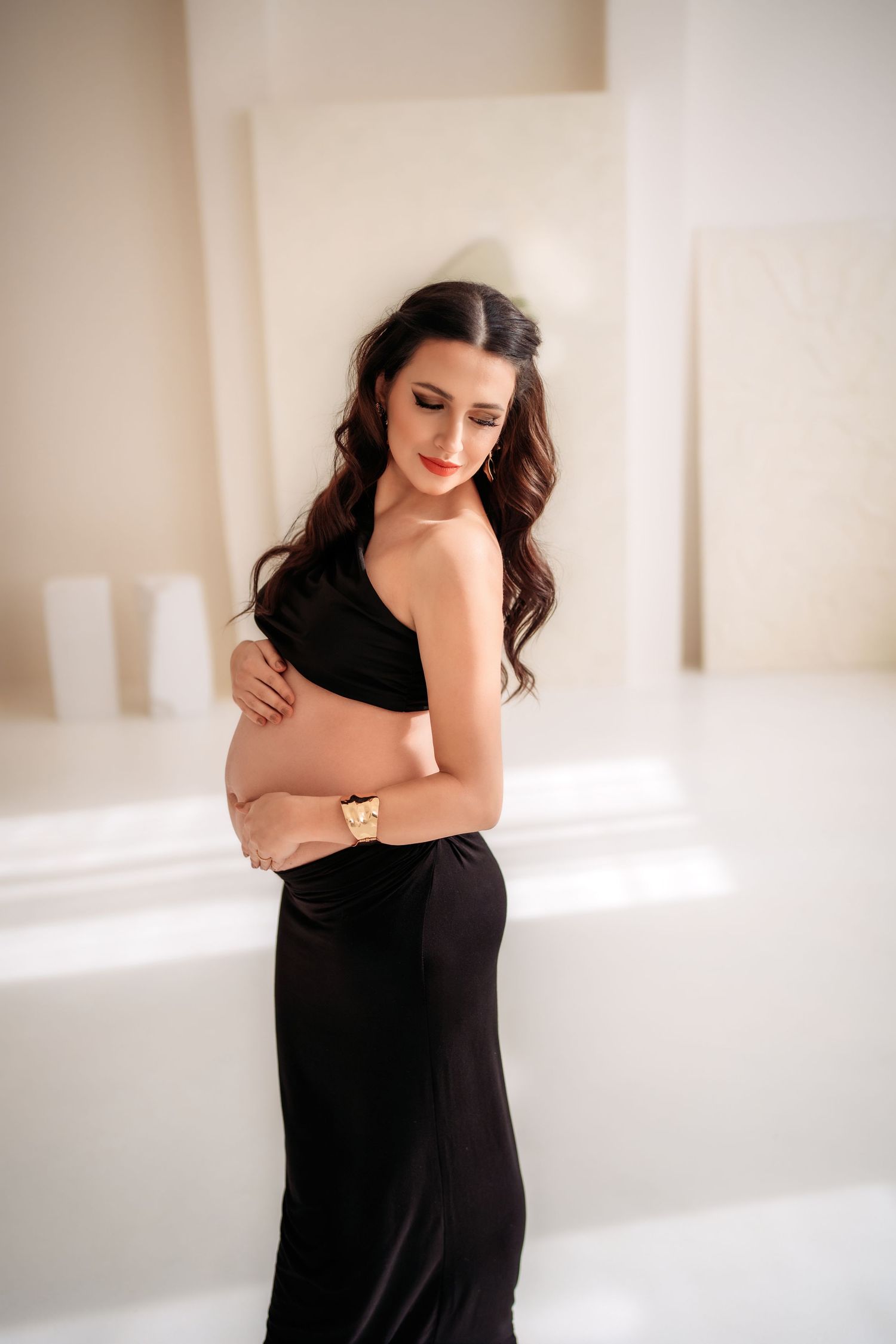 Elegant pregnant woman in black one-shoulder gown cradling baby bump, long dark wavy hair, gold watch, soft white studio.