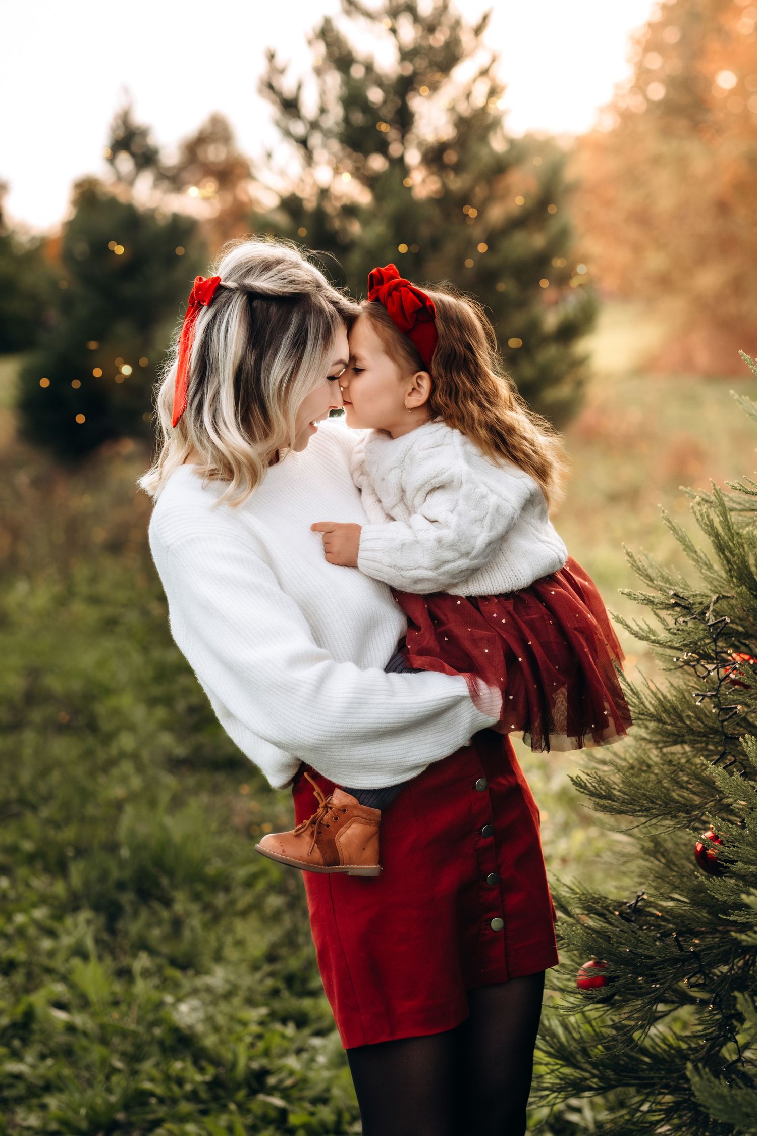 Mother and daughter in matching white sweaters and red accessories share a tender moment at a Christmas tree farm.