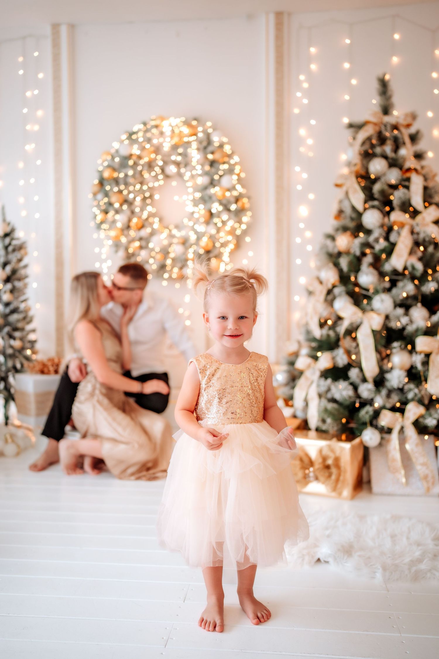 Young girl in gold dress smiles before Christmas tree, parents sharing moment behind her in festive decorated room.