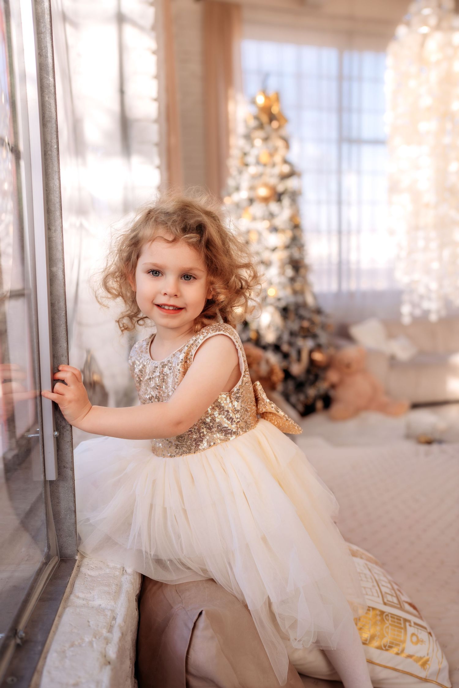 Adorable curly-haired girl in gold sequin dress poses by window with decorated Christmas tree in background.