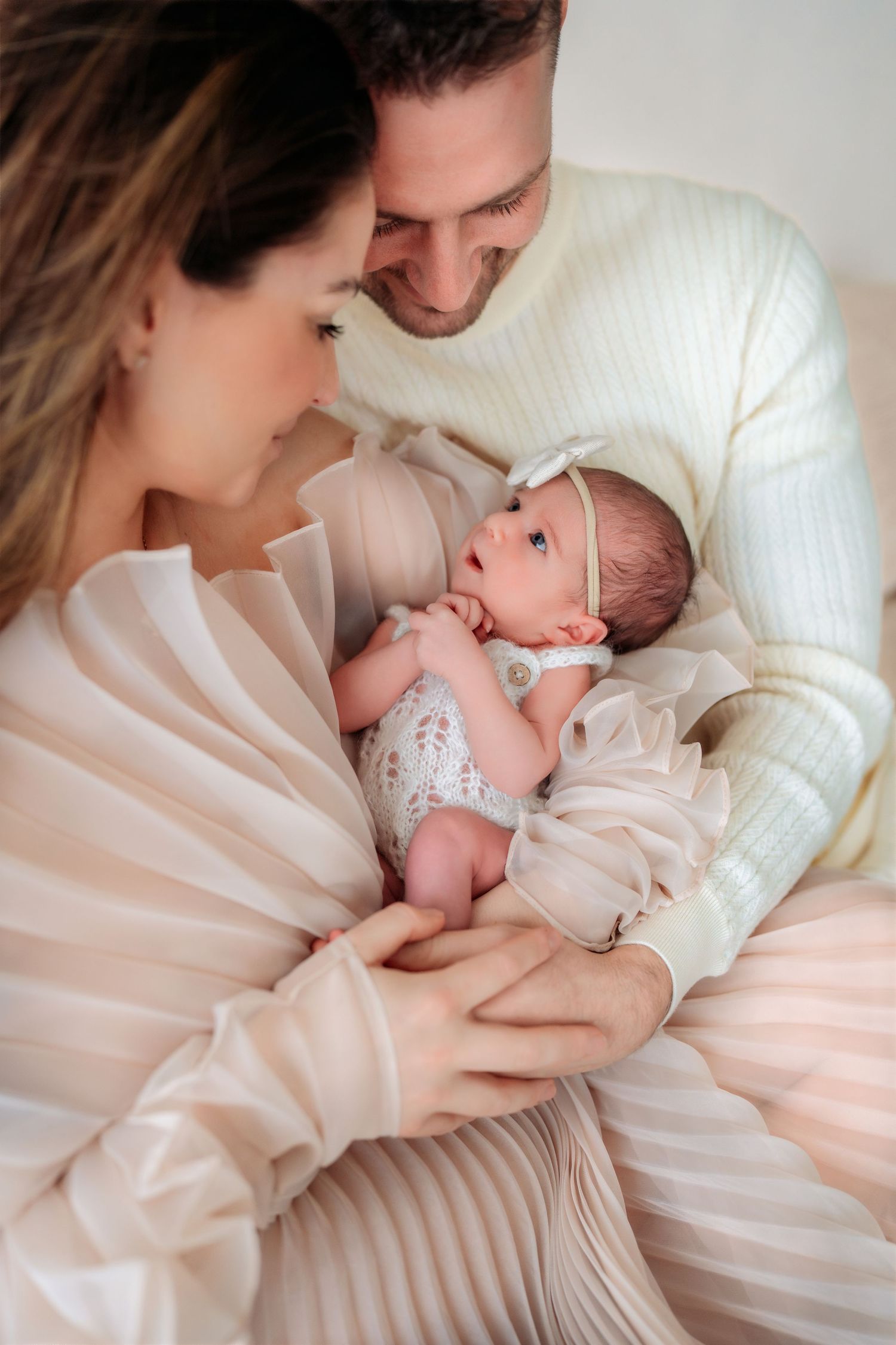 Parents in cream and pink outfits smiling at each other while cradling alert newborn baby girl.