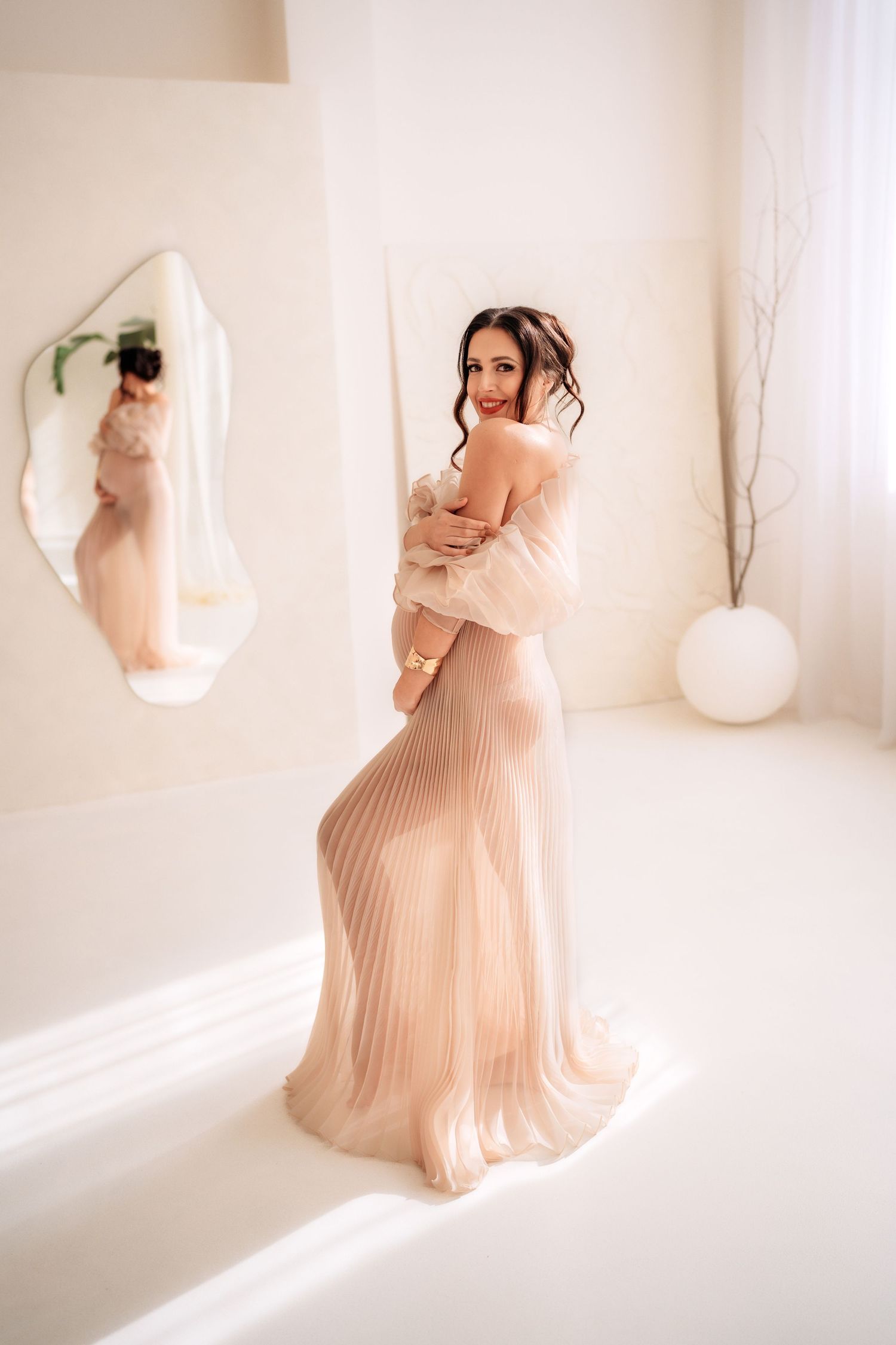Pregnant woman in elegant beige pleated maternity gown posing near mirror in white studio.