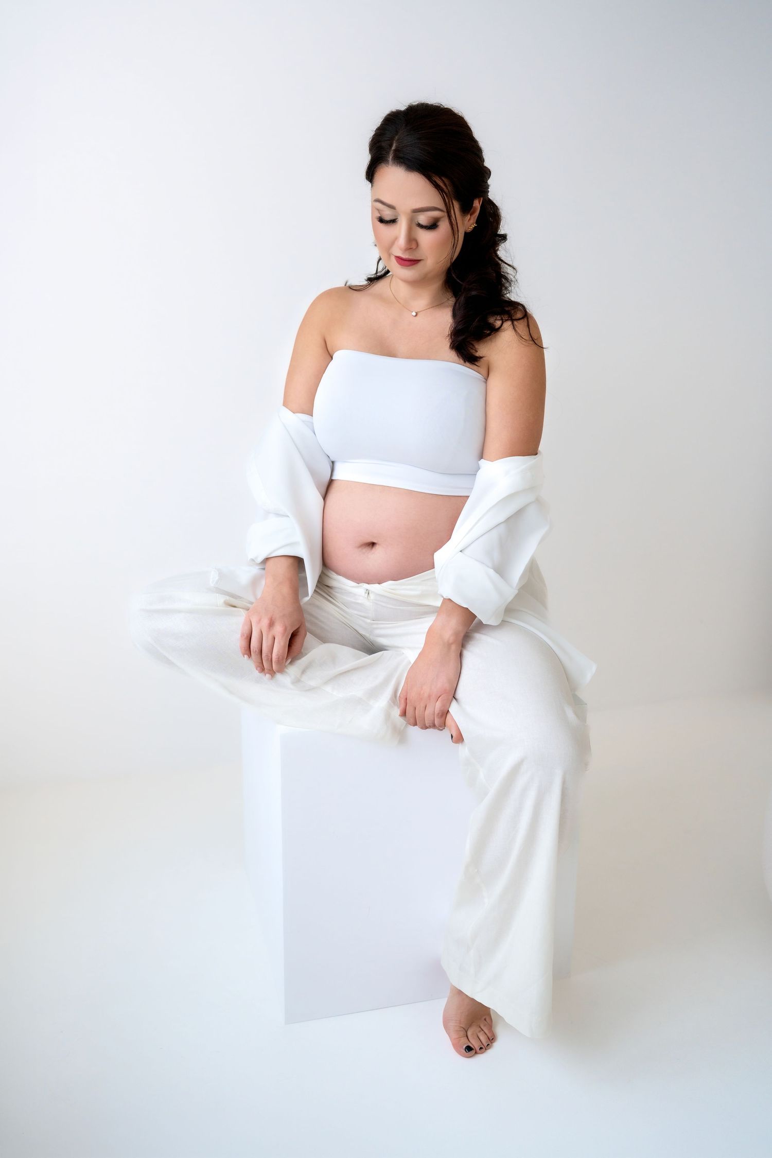 Pregnant woman in white crop top and pants sitting on cube, looking down at her baby bump in studio.