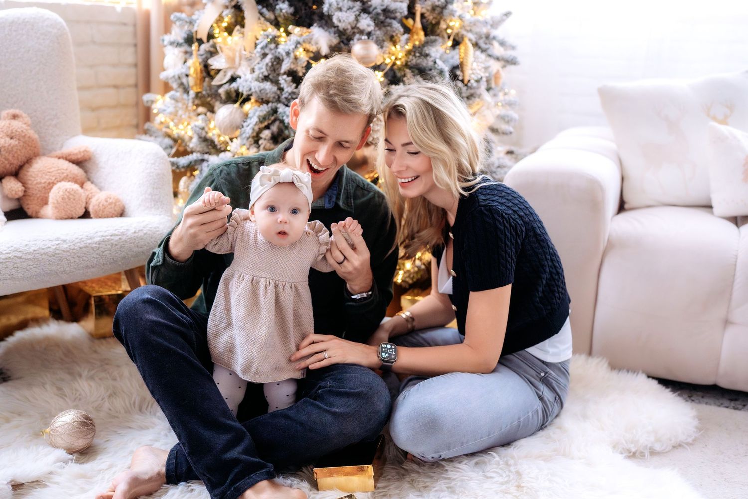 Young family with baby celebrating Christmas near decorated tree, sitting on white fur rug in cozy living room.