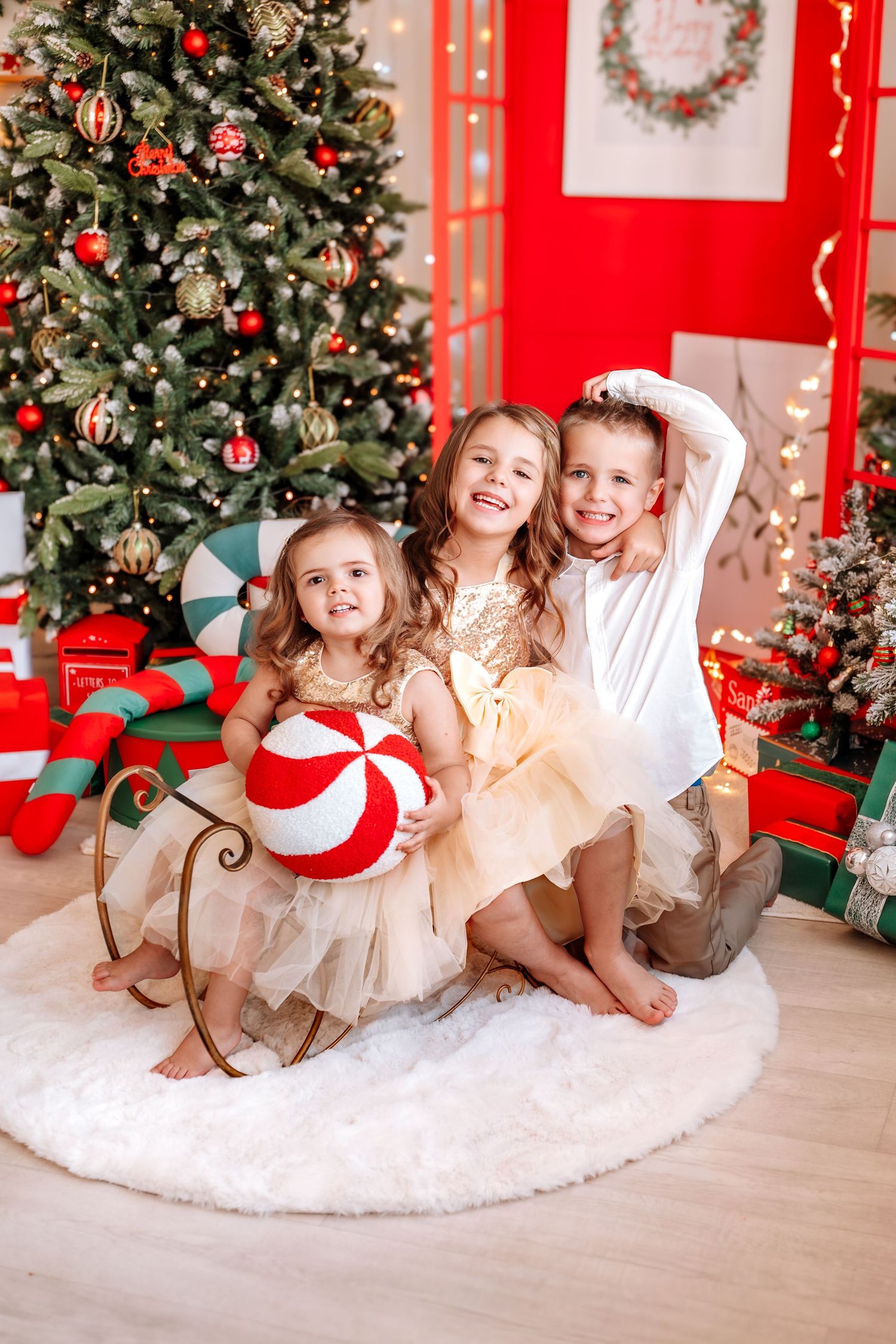 Three children pose joyfully by a decorated Christmas tree with gifts, wearing festive gold outfits and holding a candy ornament.