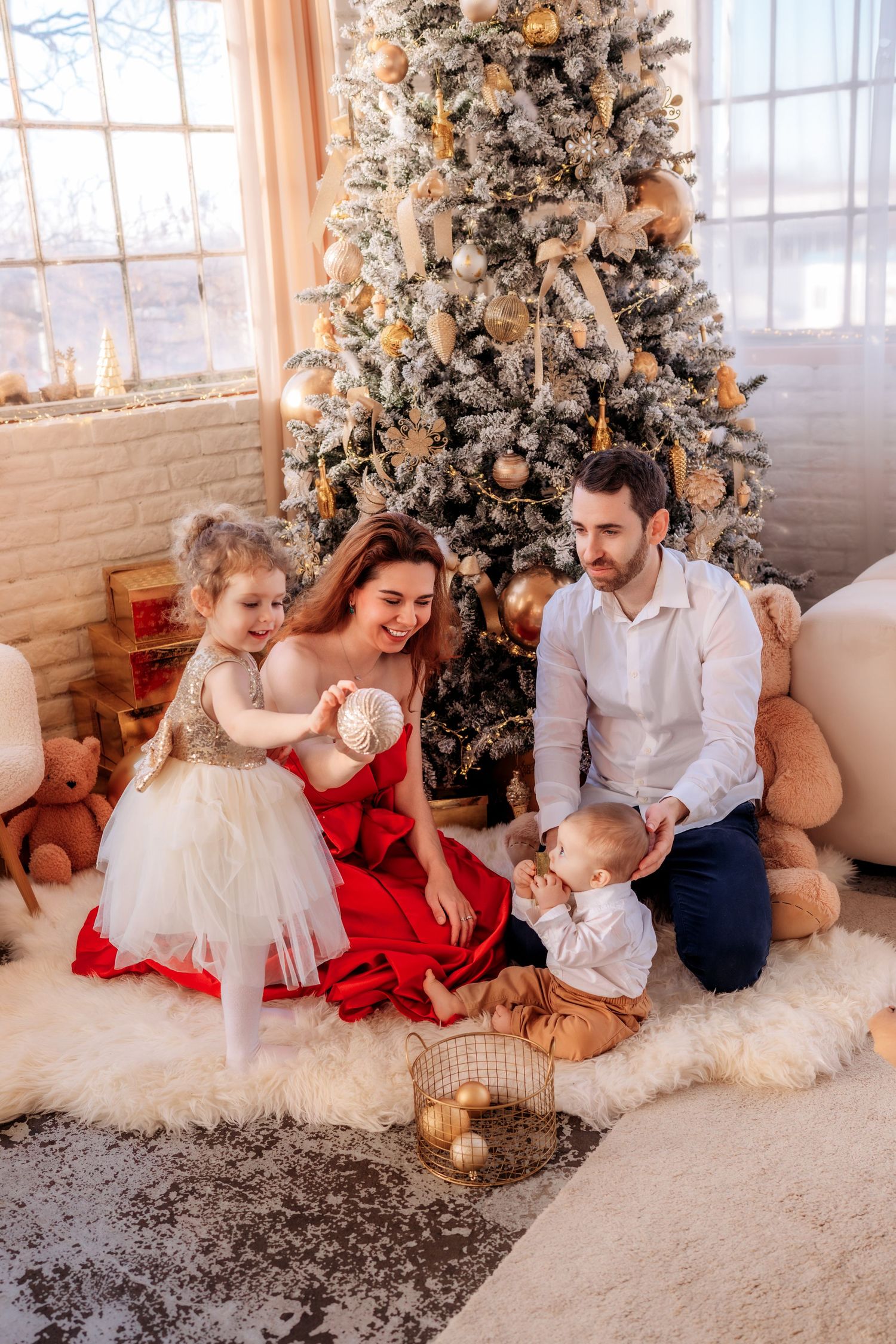 Family of four celebrating Christmas by decorated tree, parents and two children playing with ornaments on white fur rug.