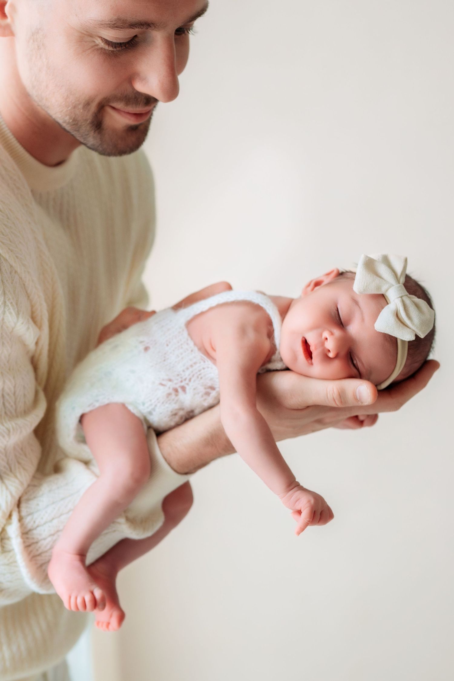 Smiling father in cream sweater carefully holding his sleeping newborn baby girl.