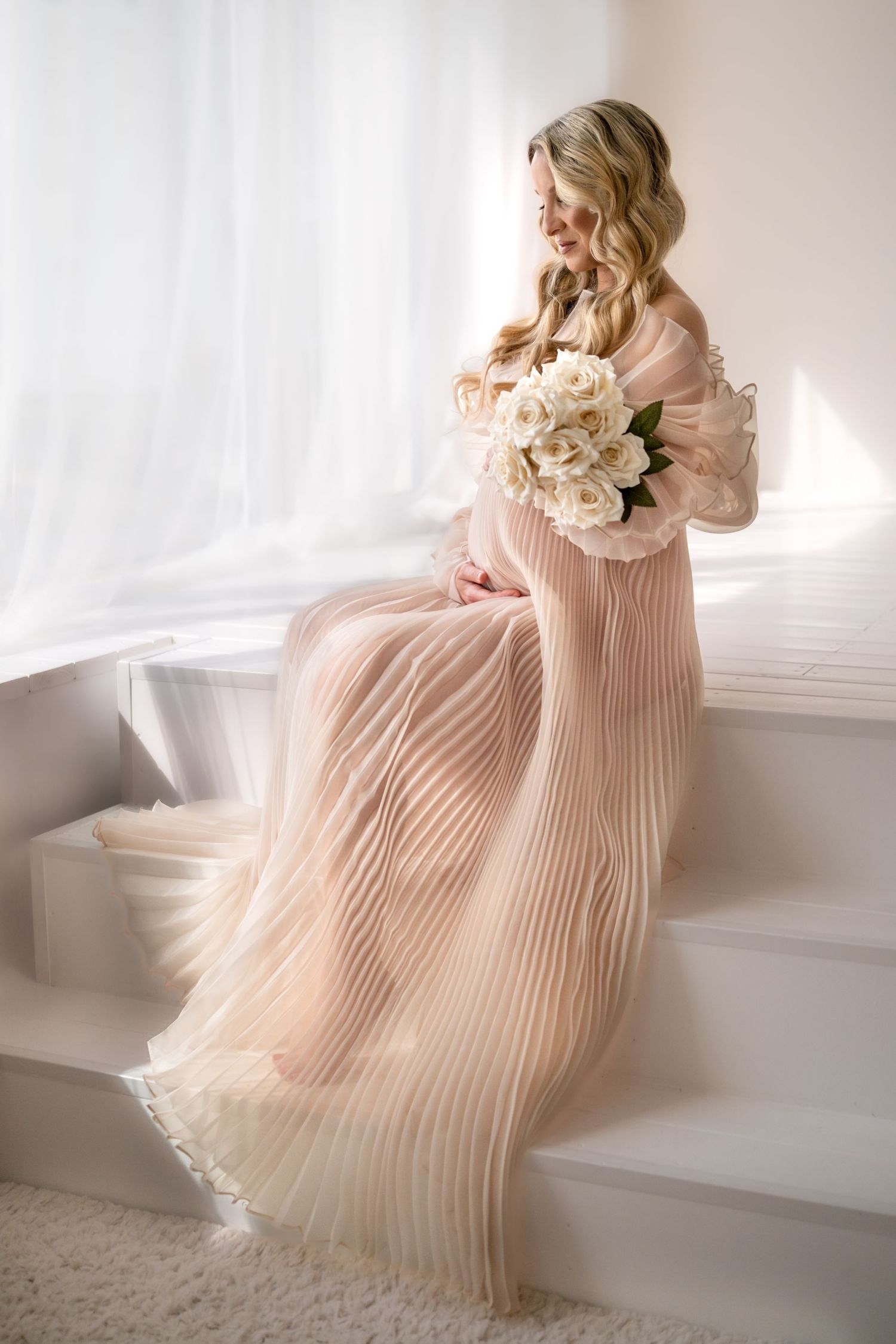Pregnant woman in blush pleated gown sitting on white steps holding white peony bouquet in sunlight.
