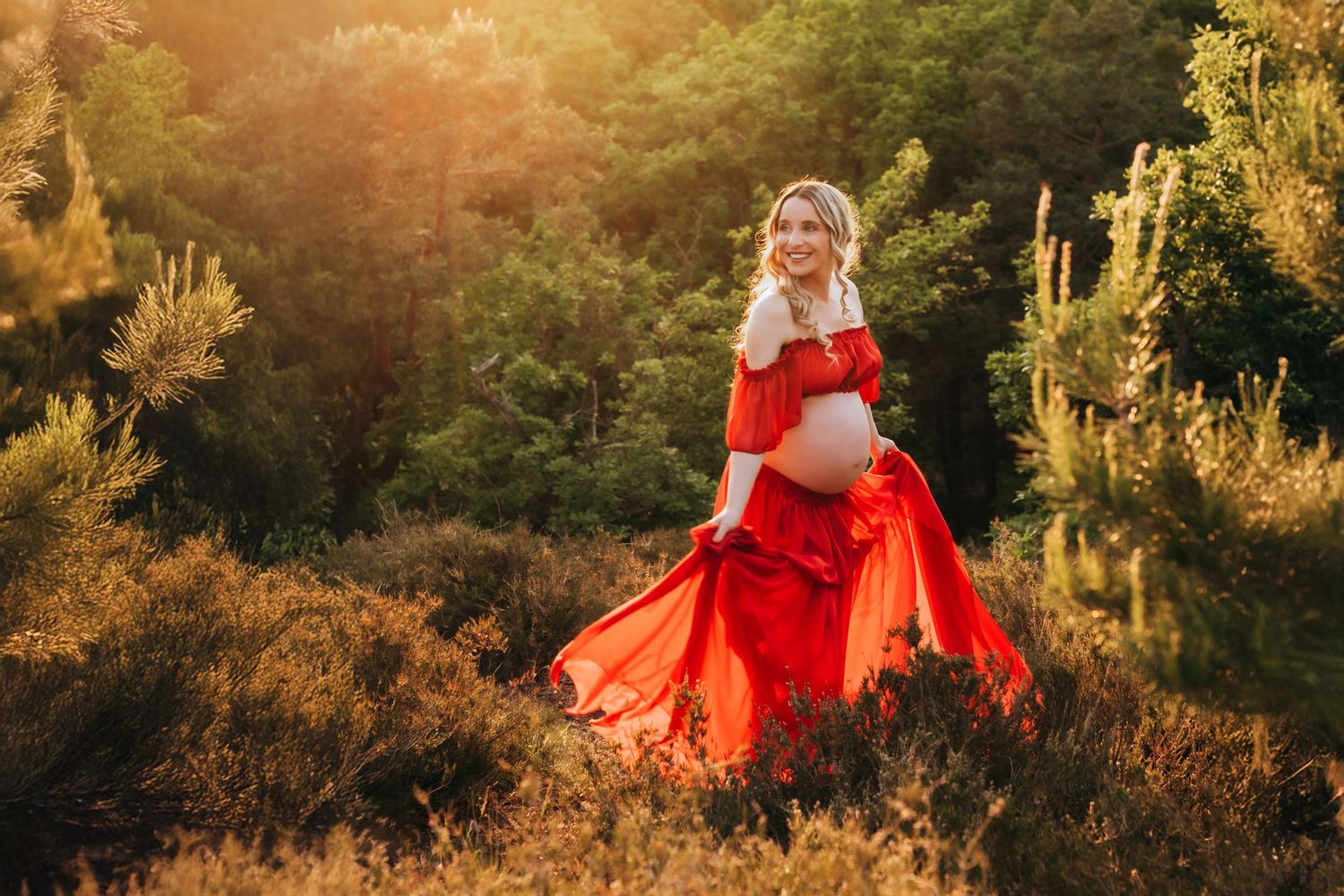 Pregnant woman in vibrant red dress standing in warm golden sunlight surrounded by green nature.