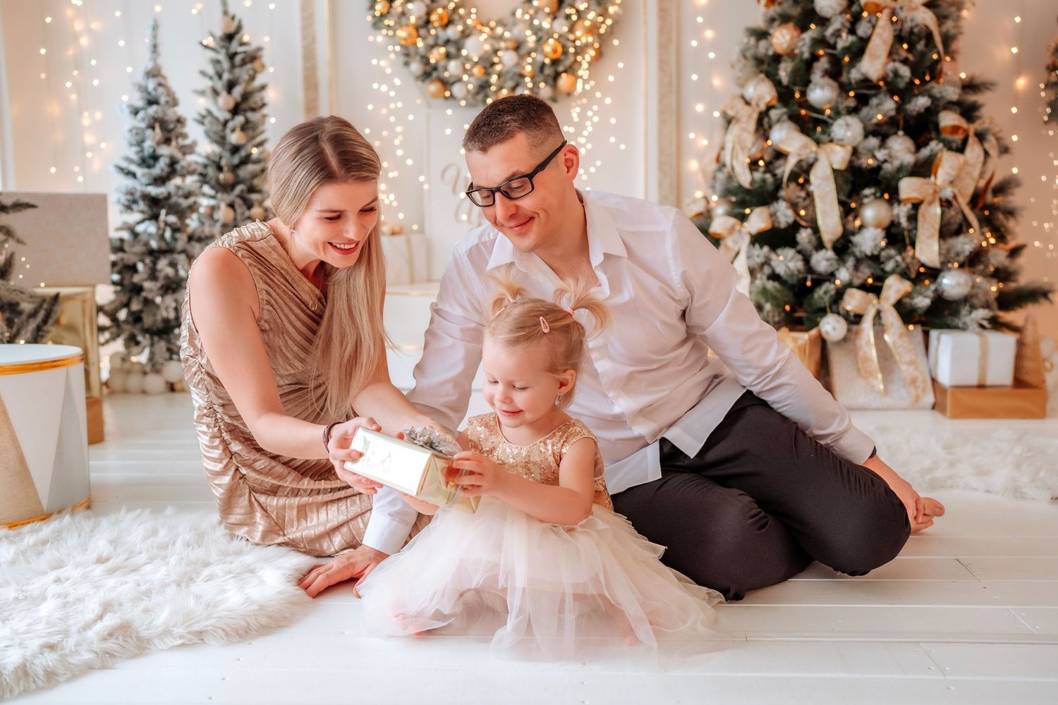 Family with young daughter opening Christmas gift near decorated tree in festive holiday living room setting.