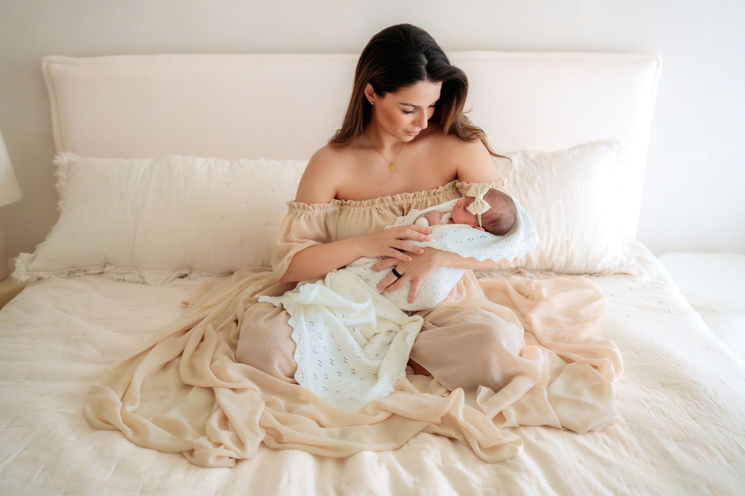 Mother in beige off-shoulder dress sitting on white bed cradling sleeping newborn wrapped in lace blanket.