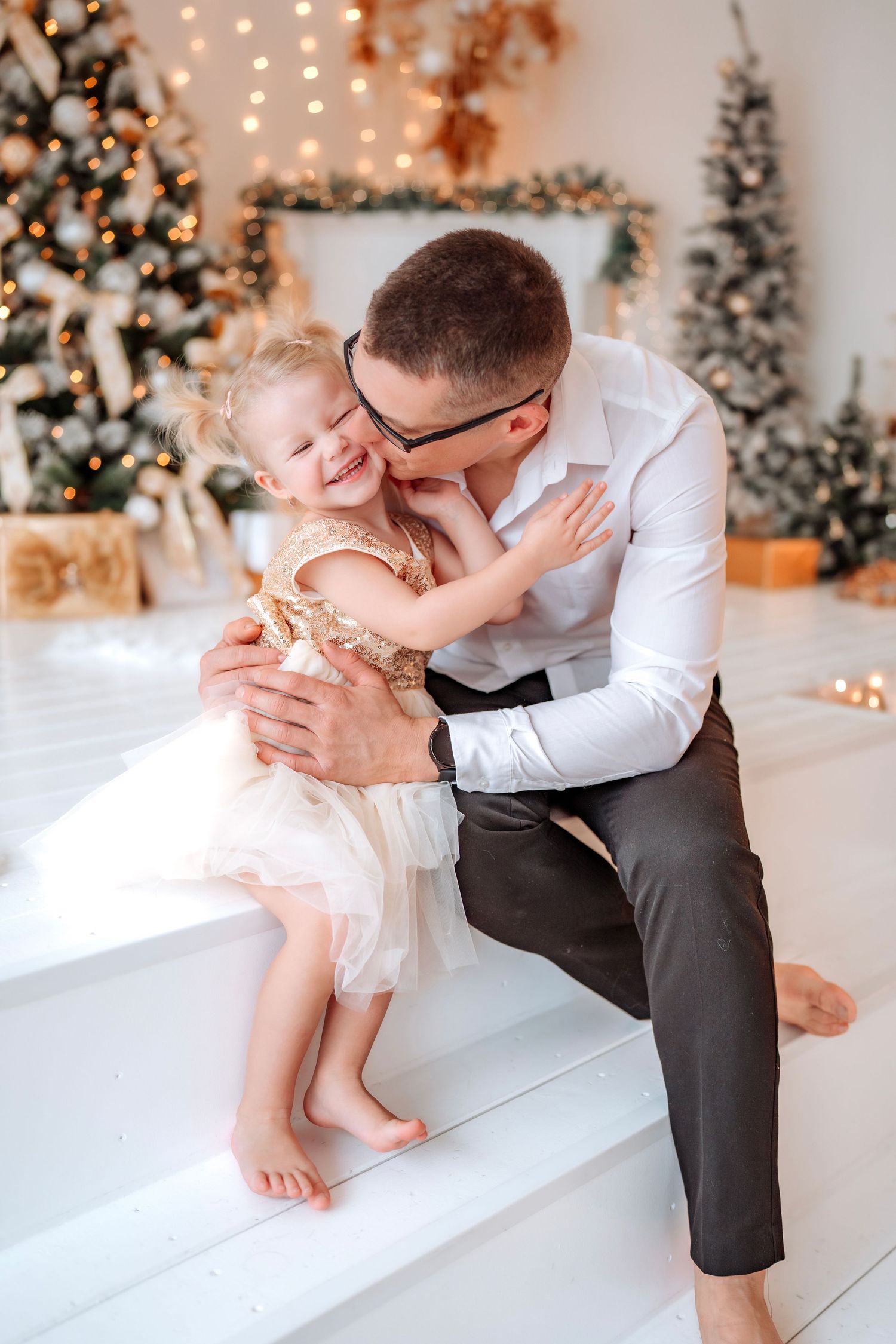 Father kissing his laughing little girl in a gold dress on white steps with Christmas trees in the background.