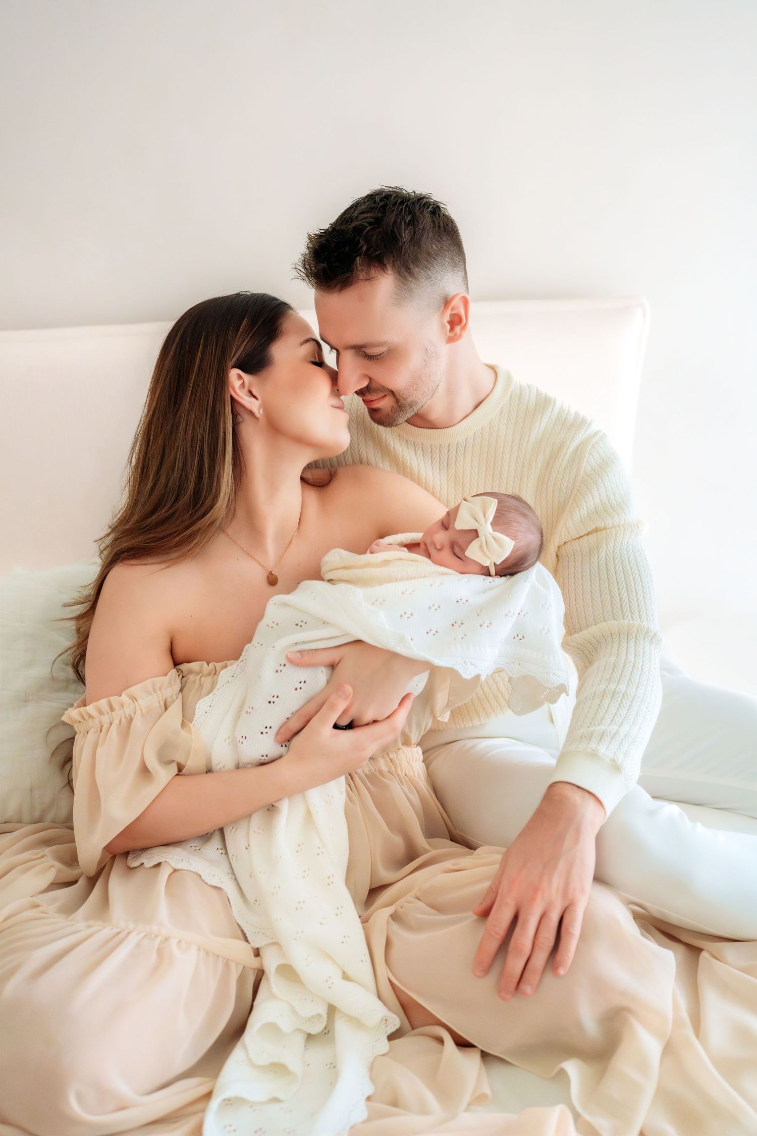 Couple in cream outfits sitting on bed touching foreheads while mother holds swaddled newborn baby.
