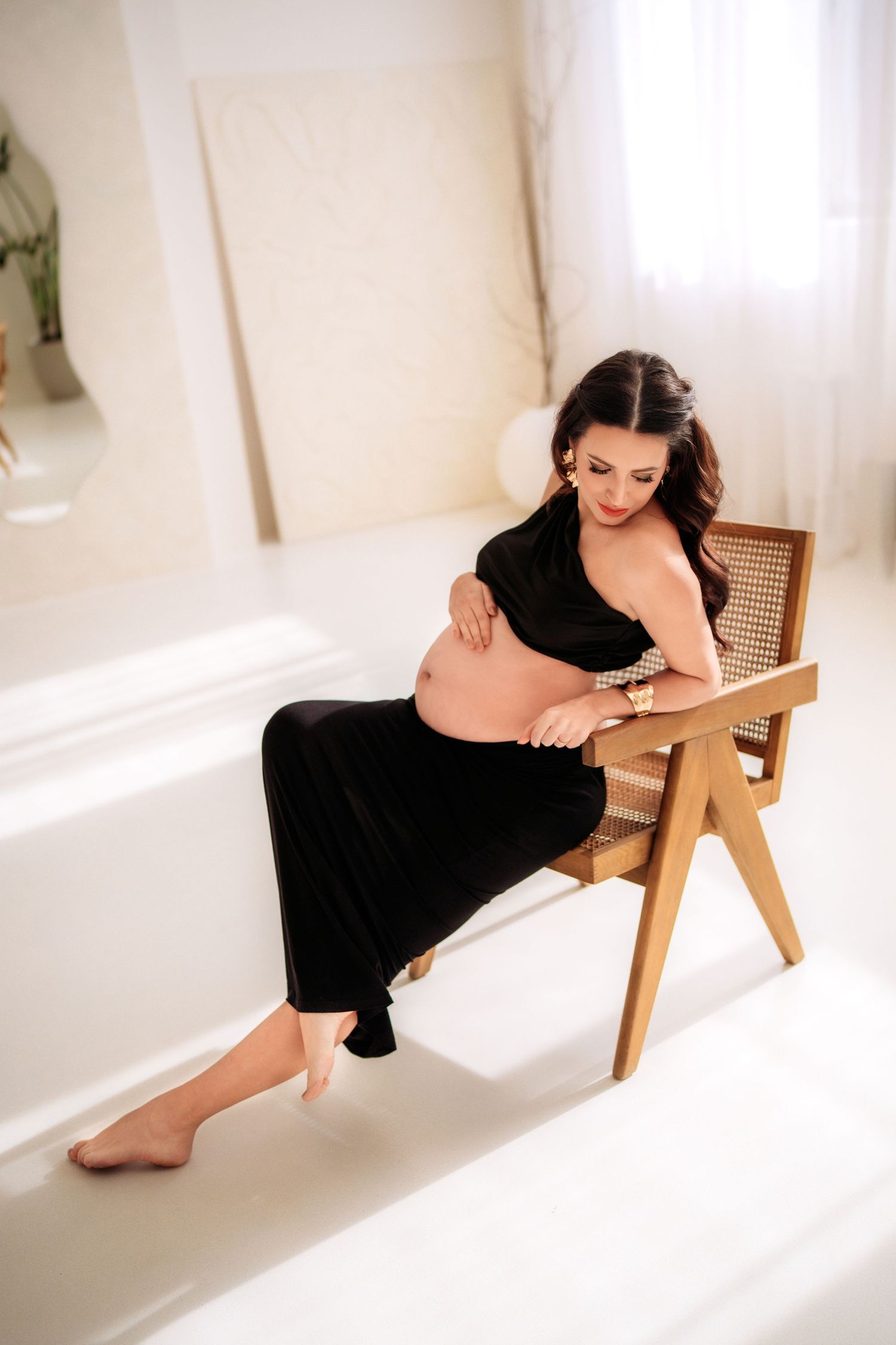 Pregnant woman in black outfit sits on wooden chair cradling her baby bump in a bright white studio.