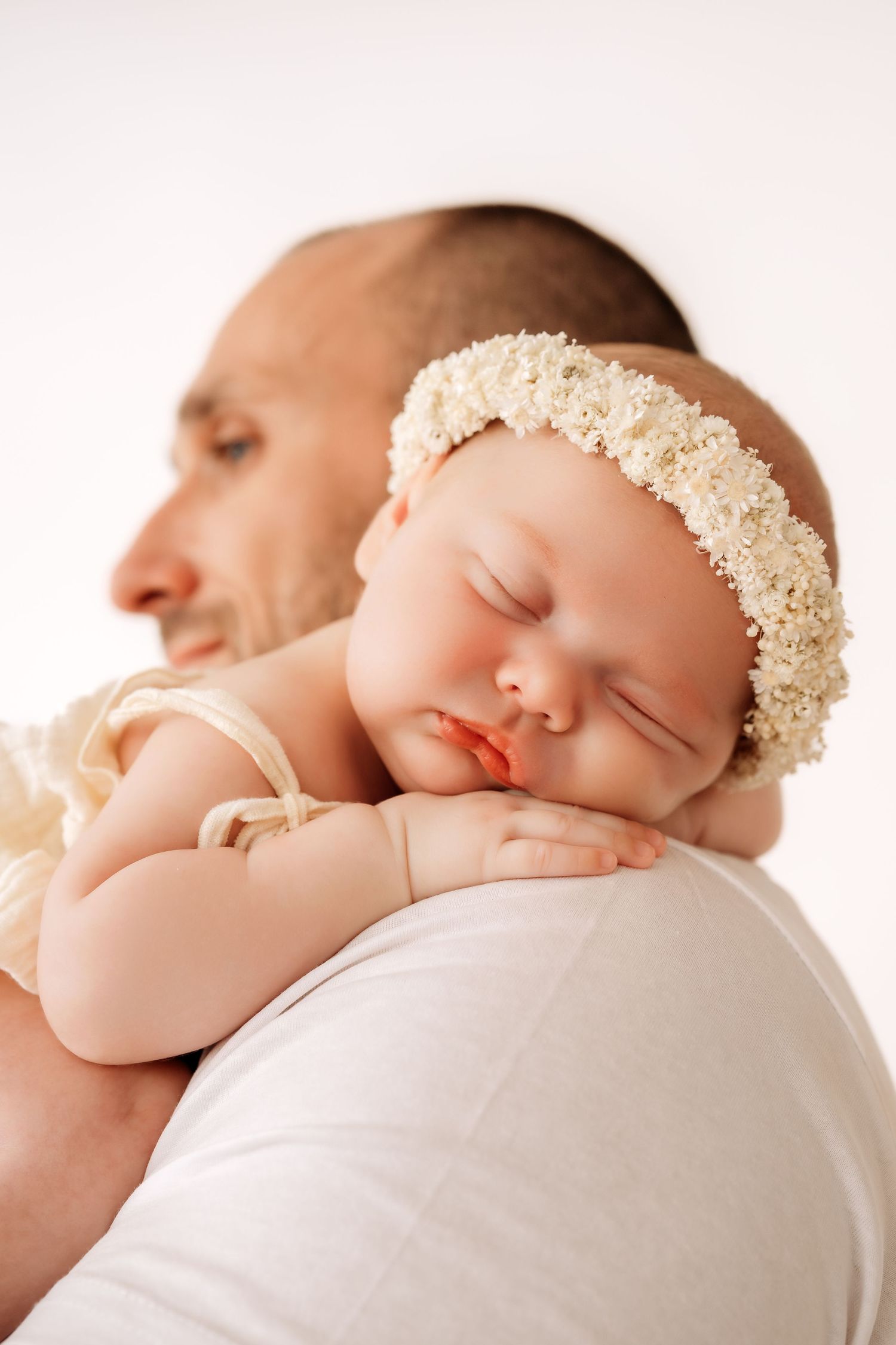 Newborn baby girl with white floral headband sleeping soundly on dads shoulder.