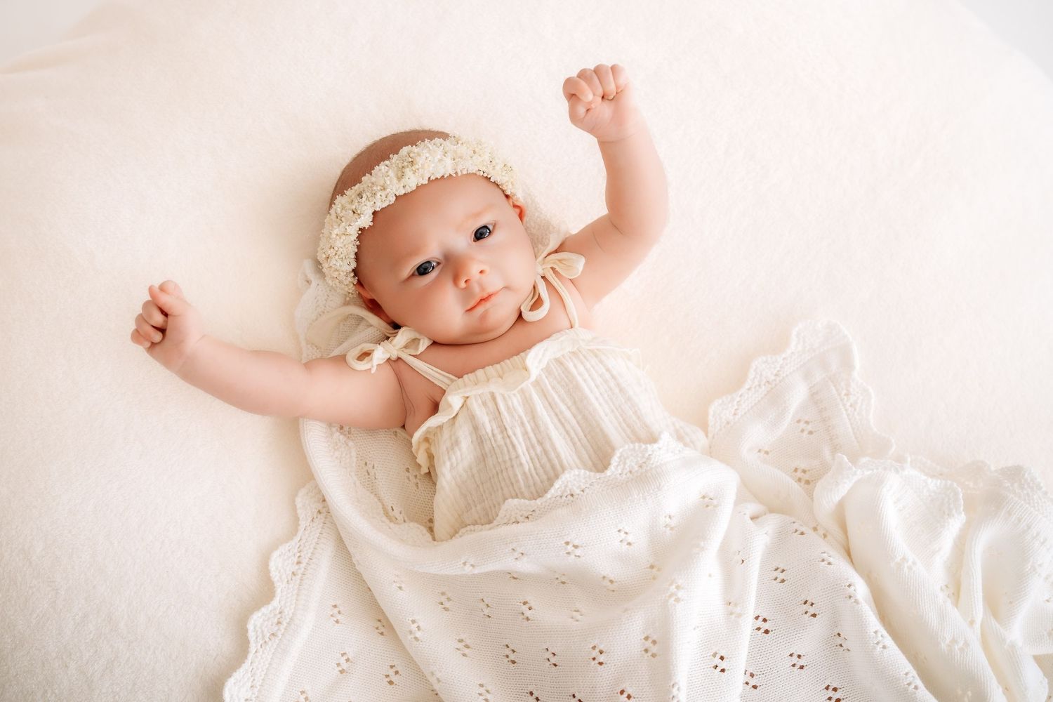Happy baby girl in white eyelet dress and lace headband stretching arms on white blanket.