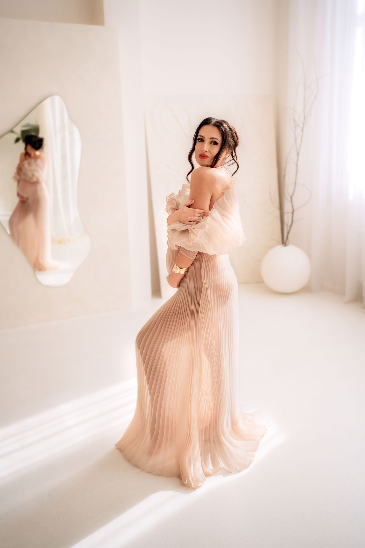 Elegant woman in sheer pleated blush gown posing gracefully in a bright white room with mirror reflection.