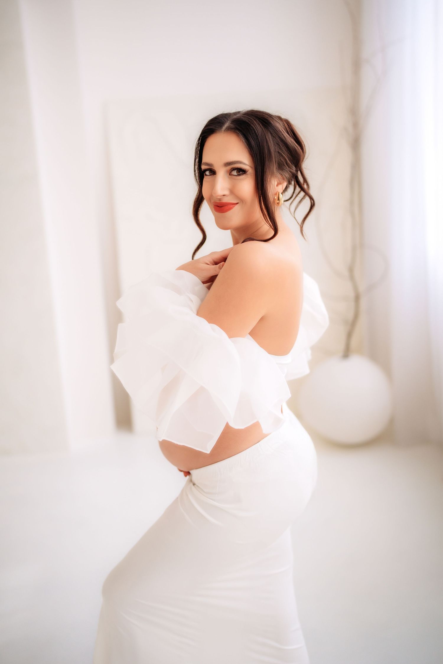 Elegant pregnant woman in white ruffled off-shoulder top and skirt posing in bright white room.