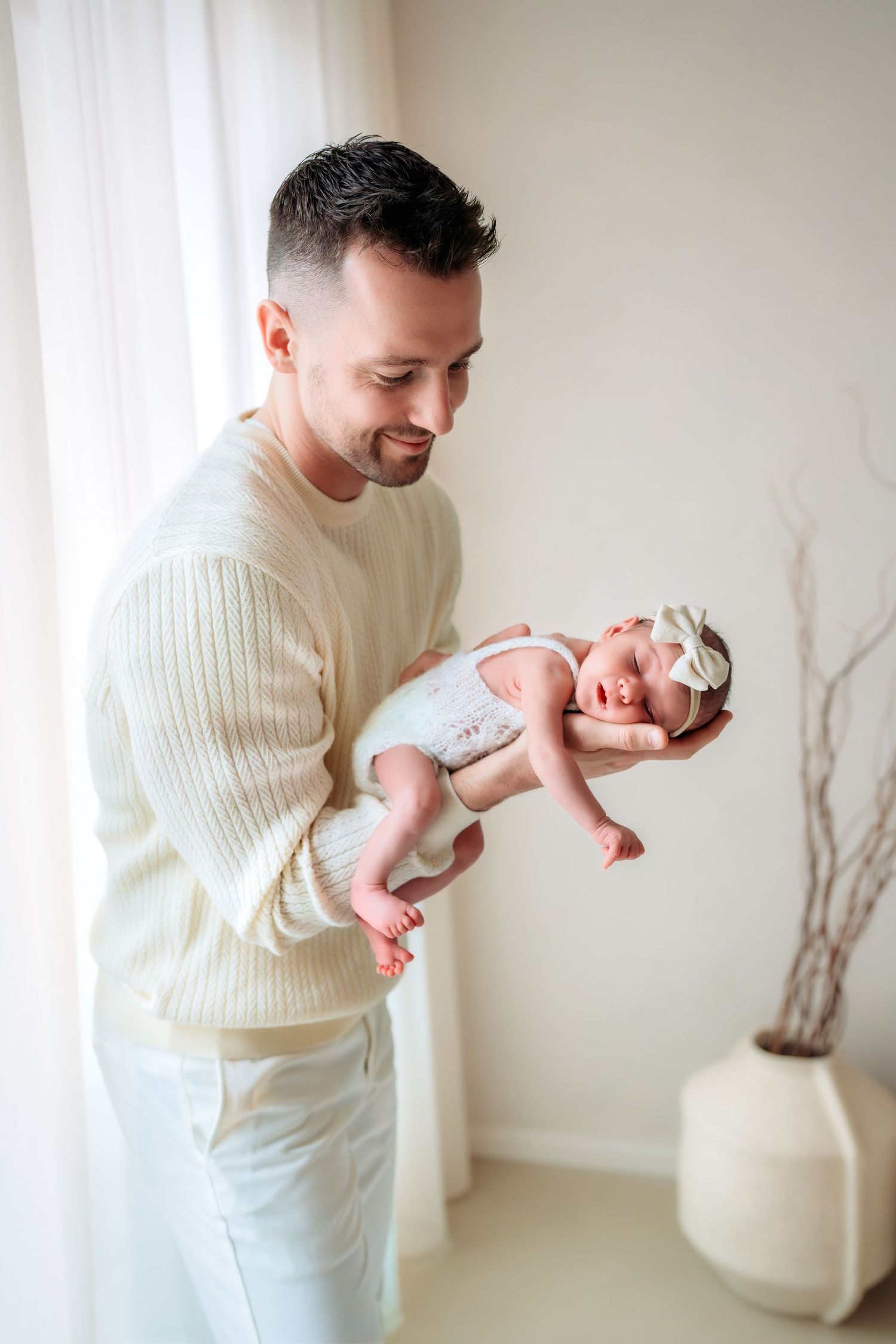 A smiling father in a cream sweater holds his newborn baby girl near a bright window at home.