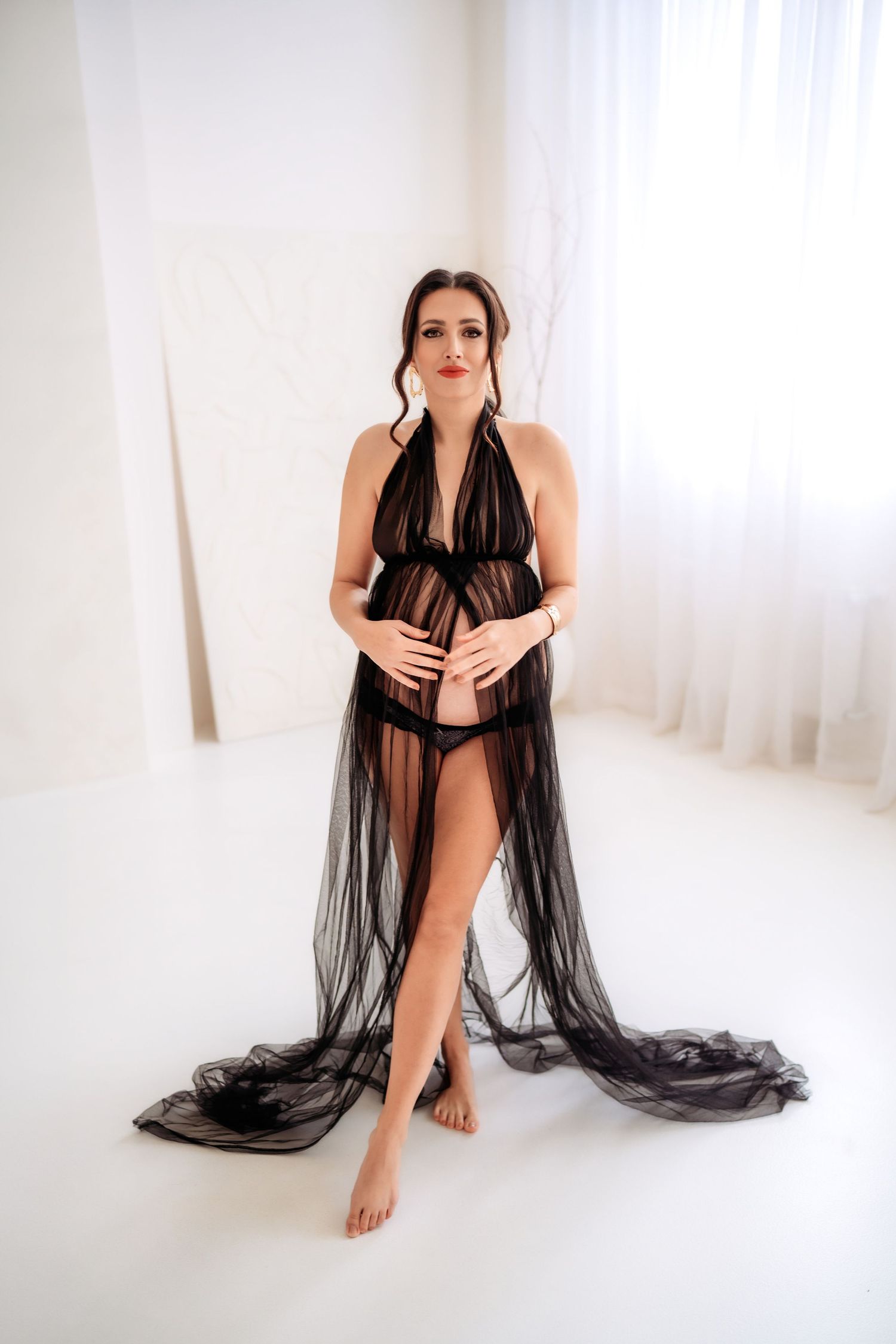 Pregnant woman in sheer black lace robe cradles baby bump while standing in white studio.