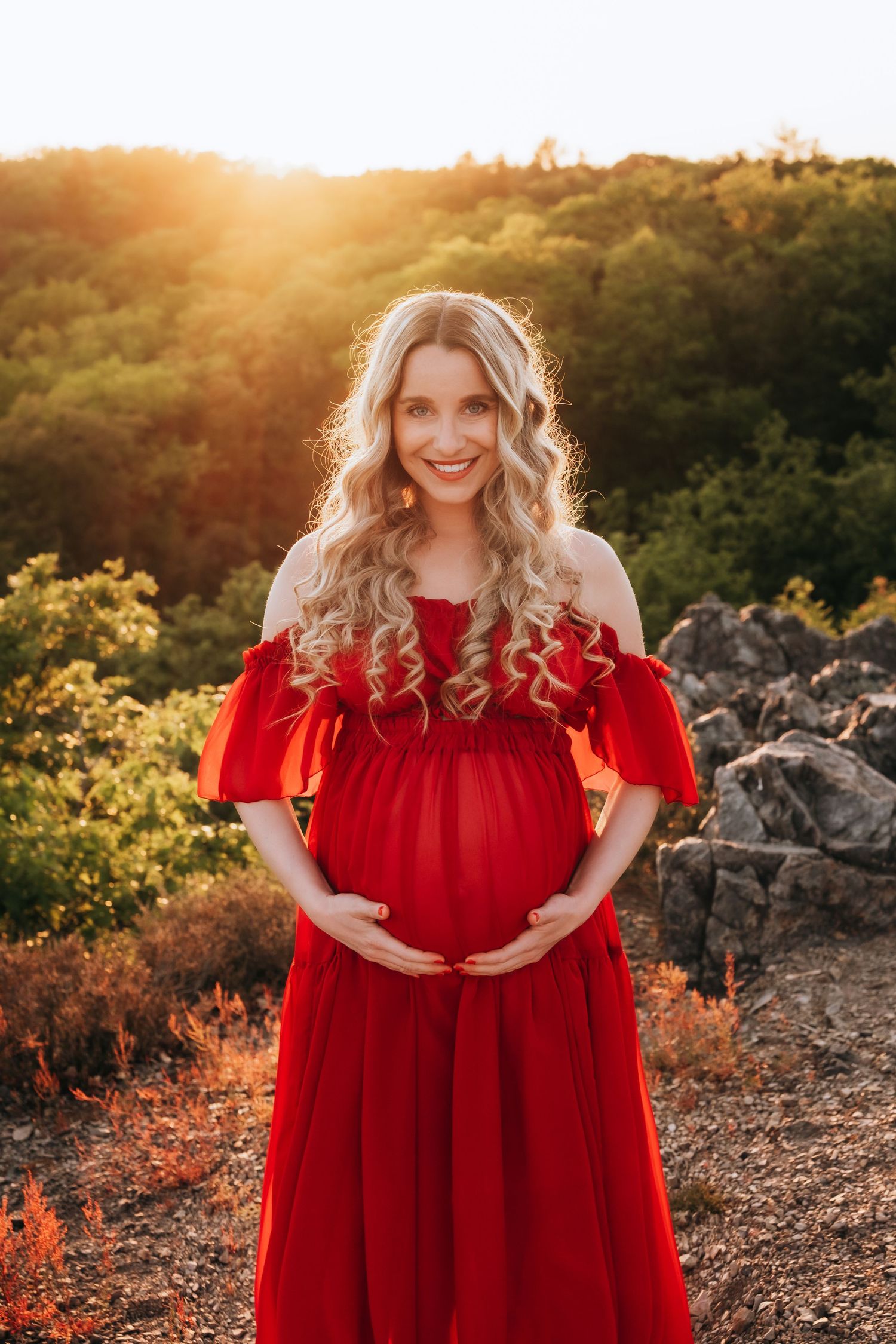 Happy pregnant blonde woman in red off-shoulder dress holding belly in warm sunset light.