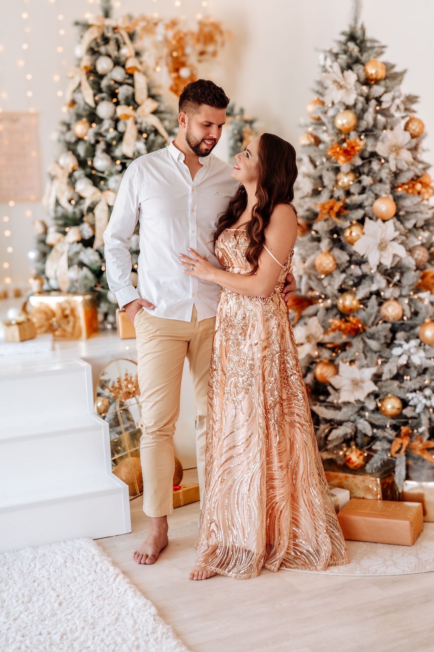 Couple in elegant attire posing by decorated Christmas trees with gold ornaments and gift boxes.