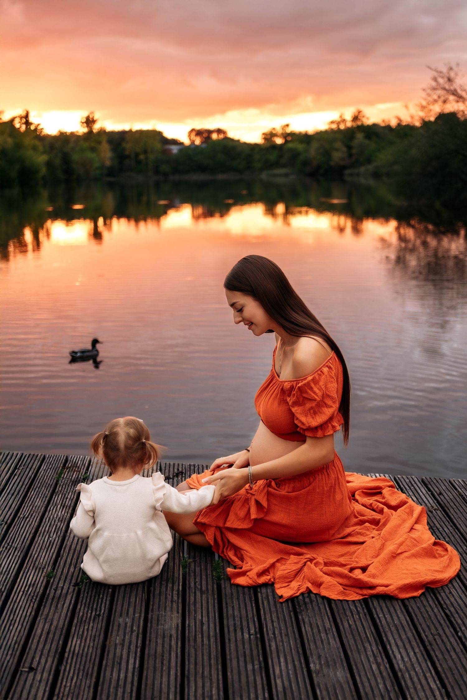 Pregnant woman in red dress sits on dock with toddler girl at sunset by a tranquil lake.