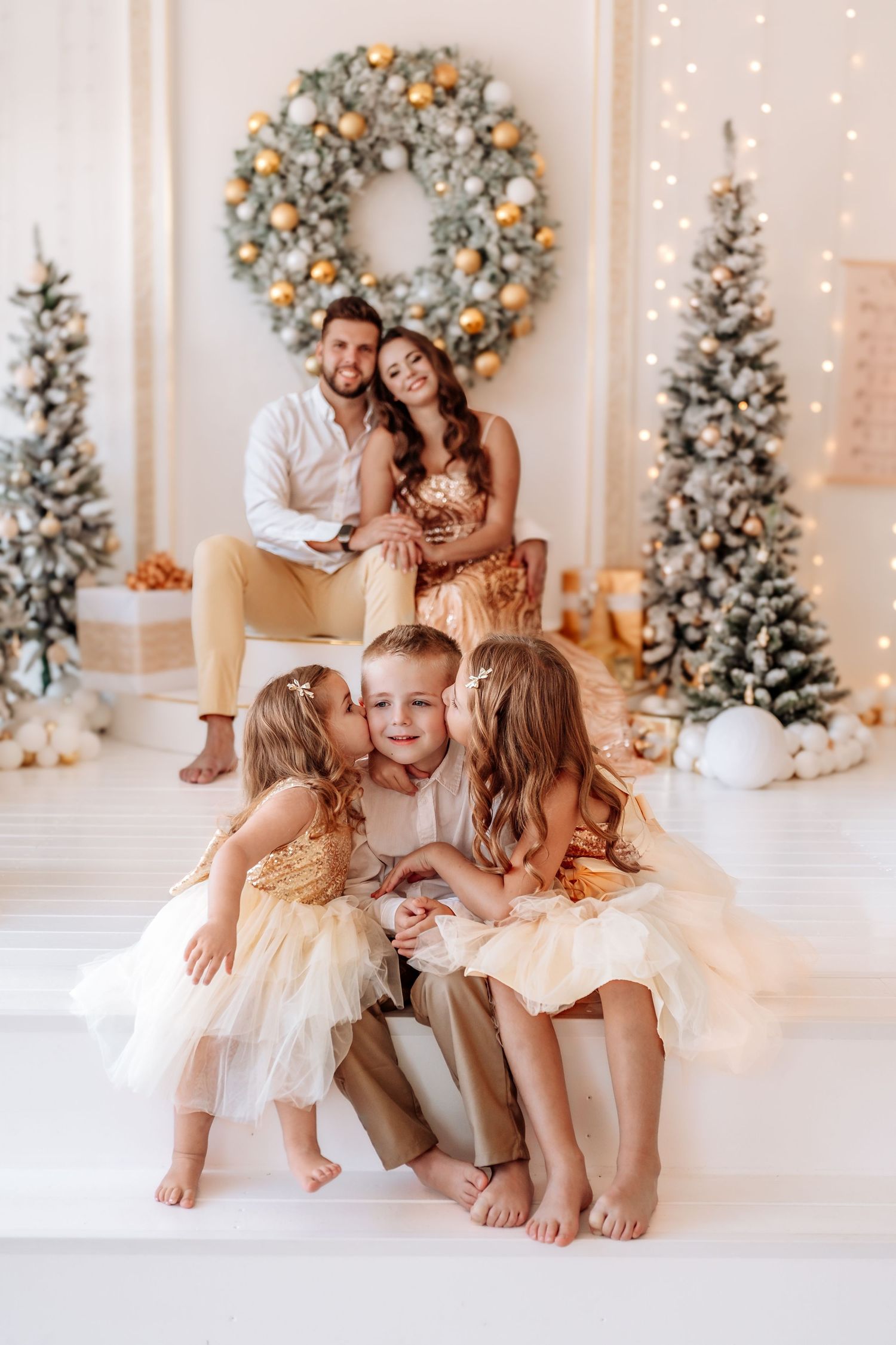 Family of five poses in festive Christmas decor with parents and three children in gold outfits near decorated trees.